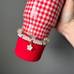 K&ouml;nnte beinhalten: Ein rot-wei&szlig; karierter Becher mit einem dekorativen Blumenband. Der untere Teil ist einfarbig rot mit einem gepr&auml;gten Design. Das Blumenband hat wei&szlig;e Perlen, Perlen und kleine Bl&uuml;ten. Eine goldene Kette ist sichtbar.
