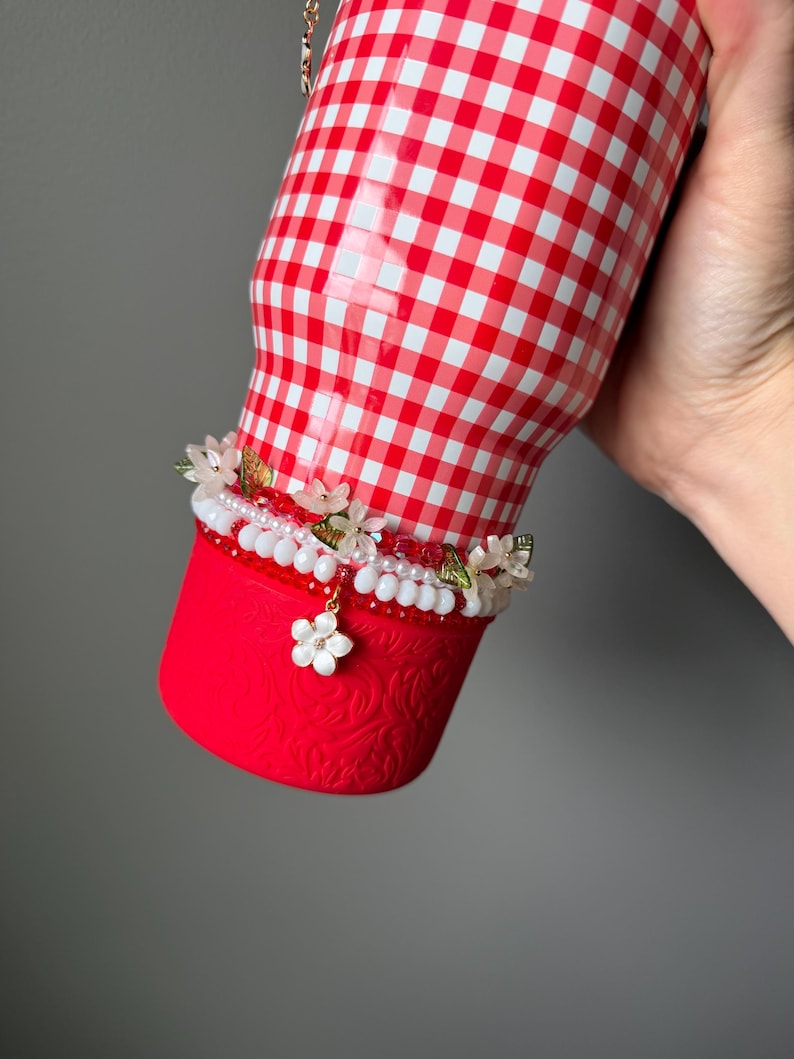K&ouml;nnte beinhalten: Ein rot-wei&szlig; karierter Becher mit einem dekorativen Blumenband. Der untere Teil ist einfarbig rot mit einem gepr&auml;gten Design. Das Blumenband hat wei&szlig;e Perlen, Perlen und kleine Bl&uuml;ten. Eine goldene Kette ist sichtbar.