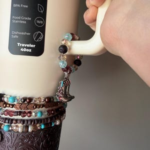 Puede incluir: Un vaso Traveler beige de 1,18L con una pulsera de cuentas decorativa y un dije de bota de vaquero plateada. El vaso tiene un dise&ntilde;o en relieve marr&oacute;n en la parte inferior e indica que es a prueba de fugas, sin BPA y apto para lavavajillas.