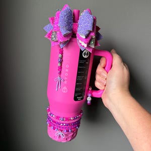 May include: A pink Hydro Jug water bottle with a handle, adorned with a large, glittery purple and pink bow. The bottle is decorated with beaded accents in shades of pink and purple.