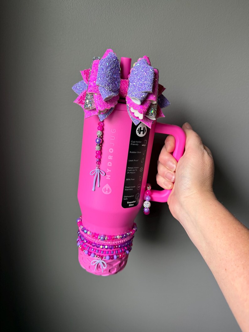 May include: A pink Hydro Jug water bottle with a handle, adorned with a large, glittery purple and pink bow. The bottle is decorated with beaded accents in shades of pink and purple.
