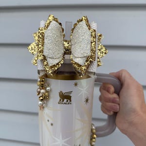 May include: A white and gold Stanley tumbler with a decorative bow and beaded accents. The bow is white with gold glitter edges. The tumbler has a handle and a straw. The design includes starbursts and the Stanley logo.