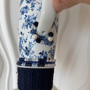 May include: A white tumbler featuring a blue floral pattern, decorated with blue and white beaded bracelets. It has a navy blue cable knit sleeve at the base. The handle is white.