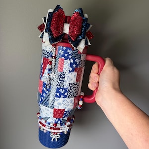 Puede incluir: Un vaso rojo, blanco y azul con un patr&oacute;n de patchwork. La tapa est&aacute; adornada con un lazo grande, brillante y de varias capas. Un dije de cuentas cuelga del costado y la base est&aacute; envuelta en una banda azul texturizada con cuentas.