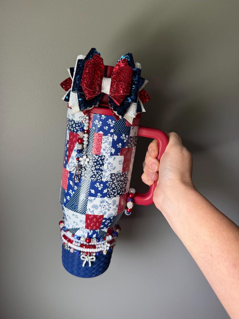 Puede incluir: Un vaso rojo, blanco y azul con un patr&oacute;n de patchwork. La tapa est&aacute; adornada con un lazo grande, brillante y de varias capas. Un dije de cuentas cuelga del costado y la base est&aacute; envuelta en una banda azul texturizada con cuentas.