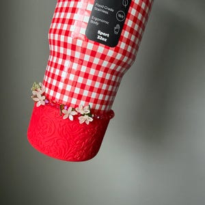 K&ouml;nnte beinhalten: Eine rot-wei&szlig; karierte Sportflasche mit roter Zierbasis. Die Flasche hat ein Fassungsverm&ouml;gen von 946 ml und eine florale Verzierung. Die Flasche besteht aus Edelstahl und ist BPA-frei.
