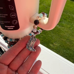 Puede incluir: Un termo blanco de 1,18L con una pulsera de abalorios y un charm de bota de vaquero plateada. La pulsera presenta cuentas turquesas, transparentes y negras. El termo es apto para lavavajillas.