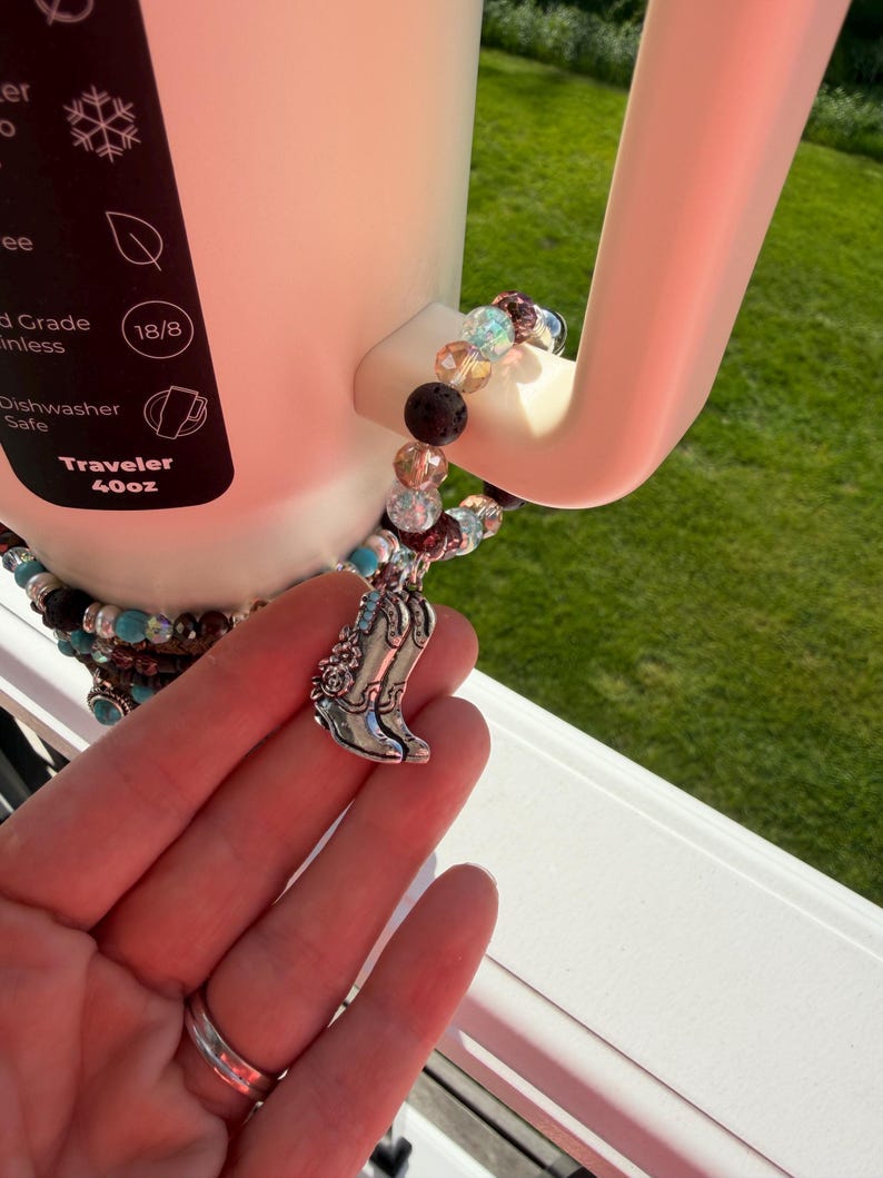 Puede incluir: Un termo blanco de 1,18L con una pulsera de abalorios y un charm de bota de vaquero plateada. La pulsera presenta cuentas turquesas, transparentes y negras. El termo es apto para lavavajillas.