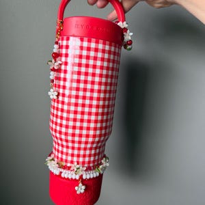 K&ouml;nnte beinhalten: Rot-wei&szlig; karierter Becher mit rotem Griff und Boden. Der Becher ist mit Perlenbl&uuml;ten in Wei&szlig;, Rot und Gr&uuml;n verziert. Das Wort "HYDRO" ist auf dem Deckel sichtbar.