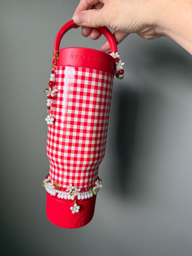K&ouml;nnte beinhalten: Rot-wei&szlig; karierter Becher mit rotem Griff und Boden. Der Becher ist mit Perlenbl&uuml;ten in Wei&szlig;, Rot und Gr&uuml;n verziert. Das Wort "HYDRO" ist auf dem Deckel sichtbar.