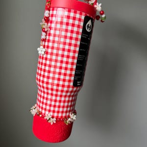 K&ouml;nnte beinhalten: Eine rot-wei&szlig; karierte Wasserflasche mit rotem Griff und Boden. Die Flasche ist mit wei&szlig;en Blumen und roten Perlen verziert. Die Flasche hat ein Fassungsverm&ouml;gen von 946 ml.