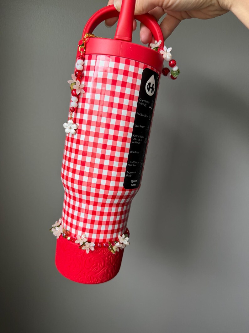 K&ouml;nnte beinhalten: Eine rot-wei&szlig; karierte Wasserflasche mit rotem Griff und Boden. Die Flasche ist mit wei&szlig;en Blumen und roten Perlen verziert. Die Flasche hat ein Fassungsverm&ouml;gen von 946 ml.