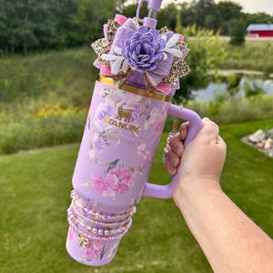 K&ouml;nnte beinhalten: Ein lilafarbener Edelstahlbecher mit Blumenmuster und einem Strohhalmdeckel. Der Becher hat einen goldenen Akzent am oberen Rand und einen goldenen Anh&auml;nger am Griff. Der Becher ist mit einer lila und goldenen Schleife mit einer wei&szlig;en Blume in der Mitte verziert.