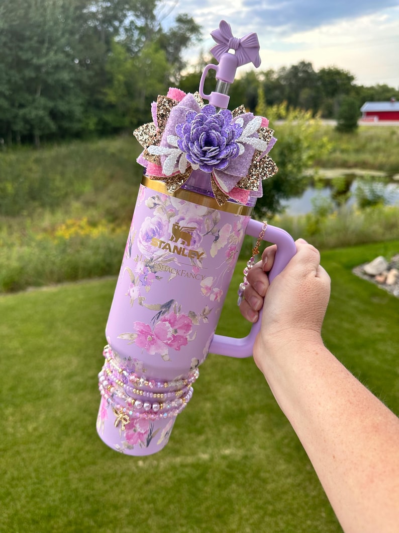 K&ouml;nnte beinhalten: Ein lilafarbener Edelstahlbecher mit Blumenmuster und einem Strohhalmdeckel. Der Becher hat einen goldenen Akzent am oberen Rand und einen goldenen Anh&auml;nger am Griff. Der Becher ist mit einer lila und goldenen Schleife mit einer wei&szlig;en Blume in der Mitte verziert.