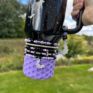 Può includere: Un bicchiere nero con manico e base viola, decorato con pipistrelli e stelle argentate glitterate. Il bicchiere è adornato con braccialetti di perline neri, viola e argento e un ciondolo a forma di pipistrello.