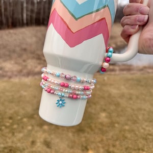 May include: A white mug with a pink, blue, and peach chevron pattern and the word "STANLEY" printed on the side. The mug is being held by a hand and has several pink, blue, and white beaded bracelets with flower charms attached.