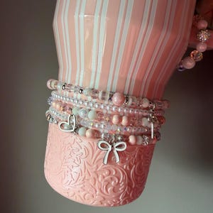 Pu&ograve; includere: Un bicchiere a righe rosa chiaro e bianco con una base testurizzata, decorato con una pila di braccialetti di perline. I braccialetti presentano varie perline rosa, bianche e trasparenti, insieme a ciondoli argentati a forma di cuore, fiocco e chiave.