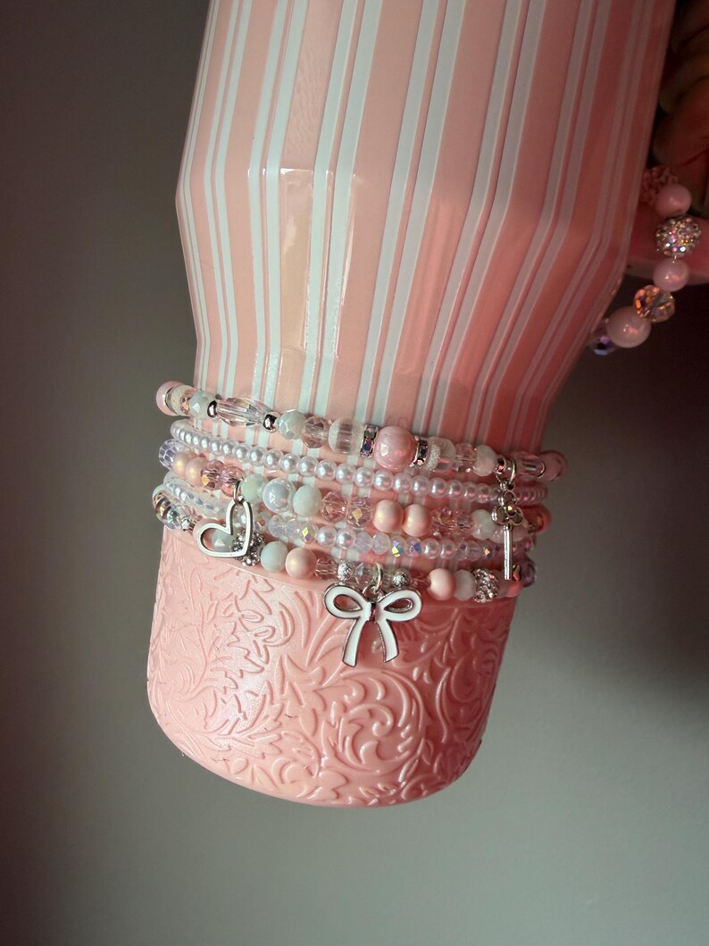 Pu&ograve; includere: Un bicchiere a righe rosa chiaro e bianco con una base testurizzata, decorato con una pila di braccialetti di perline. I braccialetti presentano varie perline rosa, bianche e trasparenti, insieme a ciondoli argentati a forma di cuore, fiocco e chiave.
