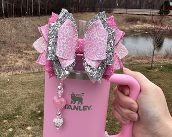 Pode incluir: Um copo térmico de aço inoxidável rosa com um laço de glitter prata e rosa, um charme de coração rosa e a palavra "STANLEY" no lado.