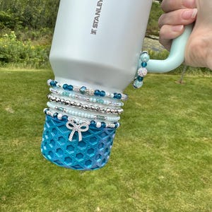 May include: A light blue Stanley water bottle with a mint green handle, adorned with multiple beaded bracelets in shades of blue, silver, and white. A decorative blue sleeve with a bow detail is at the base. The bottle is held against a backdrop of green grass.