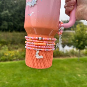 May include: A pink and orange ombre tumbler with a handle, decorated with pink and orange beaded bracelets. Silver bat charms hang from the handle and bracelets. The bottom of the tumbler has an orange textured sleeve.