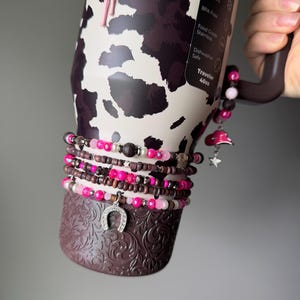 May include: A 40oz stainless steel tumbler with a cow print pattern, featuring a dark brown base with embossed floral details. The tumbler is adorned with multiple beaded bracelets in shades of pink, brown, and white, including charms shaped like a horseshoe, a pink cowboy hat, and a star.