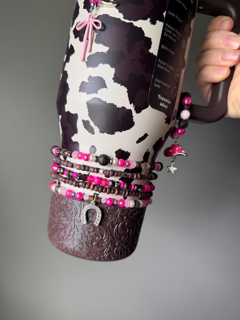 May include: A 40oz stainless steel tumbler with a cow print pattern, featuring a dark brown base with embossed floral details. The tumbler is adorned with multiple beaded bracelets in shades of pink, brown, and white, including charms shaped like a horseshoe, a pink cowboy hat, and a star.