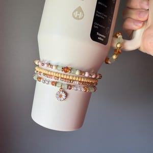 May include: A set of four beaded bracelets featuring pastel pink, pale green, and amber-colored beads, with one bracelet showcasing a white daisy charm. The bracelets are wrapped around a cream-colored 40oz stainless steel tumbler with a handle.