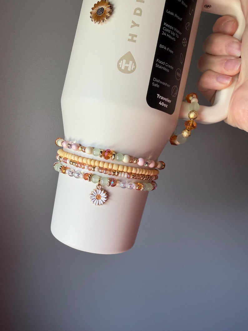 May include: A set of four beaded bracelets featuring pastel pink, pale green, and amber-colored beads, with one bracelet showcasing a white daisy charm. The bracelets are wrapped around a cream-colored 40oz stainless steel tumbler with a handle.