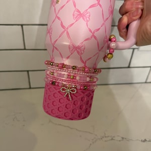 May include: Pink insulated tumbler with a bow pattern and the text "LOVESHACKFANCY". The tumbler is adorned with a gold cross charm and multiple beaded bracelets in shades of pink and gold. A pink decorative sleeve with a bow is at the bottom.