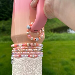 May include: A pink and peach ombre tumbler with a pink handle, adorned with beaded bracelets in shades of pink, peach, and silver. The bottom of the tumbler features a white, embossed floral design.