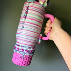 Op de afbeelding: Een roestvrijstalen beker met horizontale strepen in pastelroze, paars en turquoise. De beker heeft een roze handvat, een glinsterende veelkleurige strik aan de bovenkant en een roze basis met textuur. Versierd met kralenbedels en een hartje.