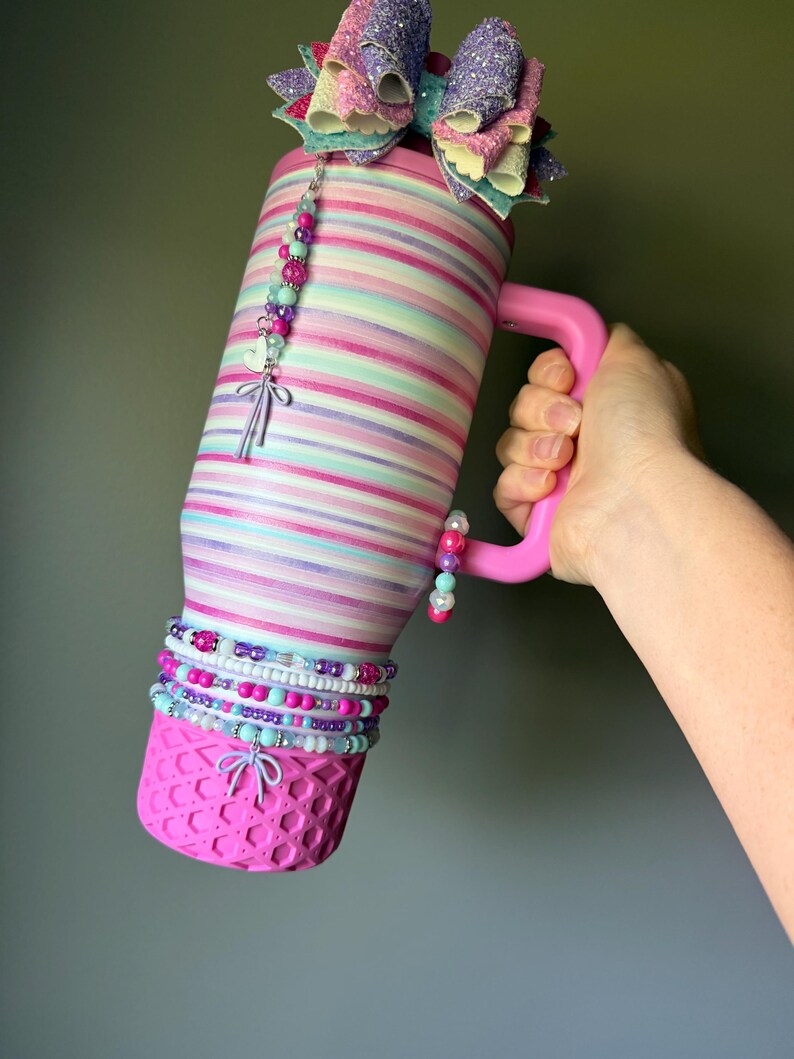 Op de afbeelding: Een roestvrijstalen beker met horizontale strepen in pastelroze, paars en turquoise. De beker heeft een roze handvat, een glinsterende veelkleurige strik aan de bovenkant en een roze basis met textuur. Versierd met kralenbedels en een hartje.