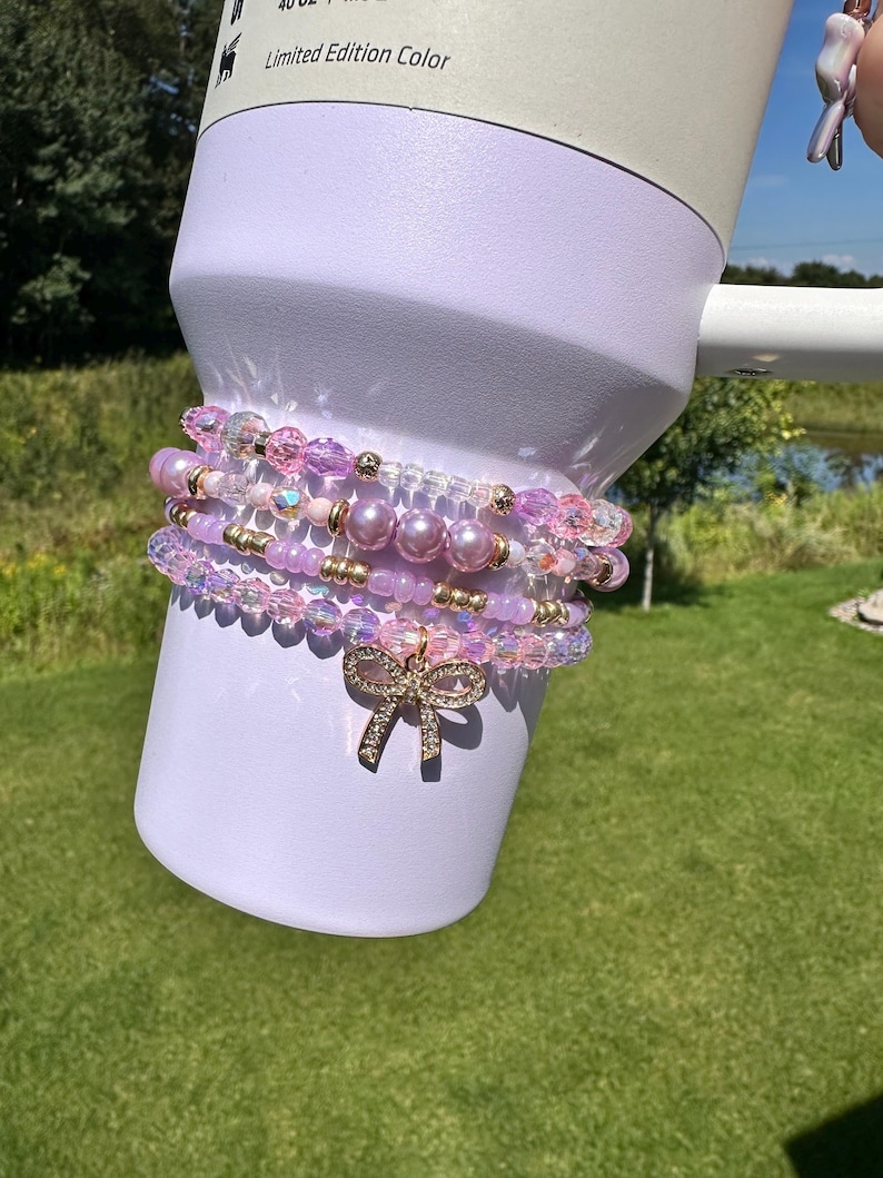 K&ouml;nnte beinhalten: Ein Set aus vier rosafarbenen und schillernden Perlenarmb&auml;ndern mit einem goldenen Schleifenanh&auml;nger. Die Armb&auml;nder sind auf einem wei&szlig;en Becher gestapelt, auf dem der Text "Limited Edition Color" steht.