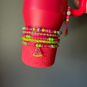May include: A bright pink tumbler with a textured base and a handle, adorned with multiple stacked beaded bracelets in neon green and pink. The bracelets feature various round beads, faceted crystals, and a dangling watermelon slice charm.
