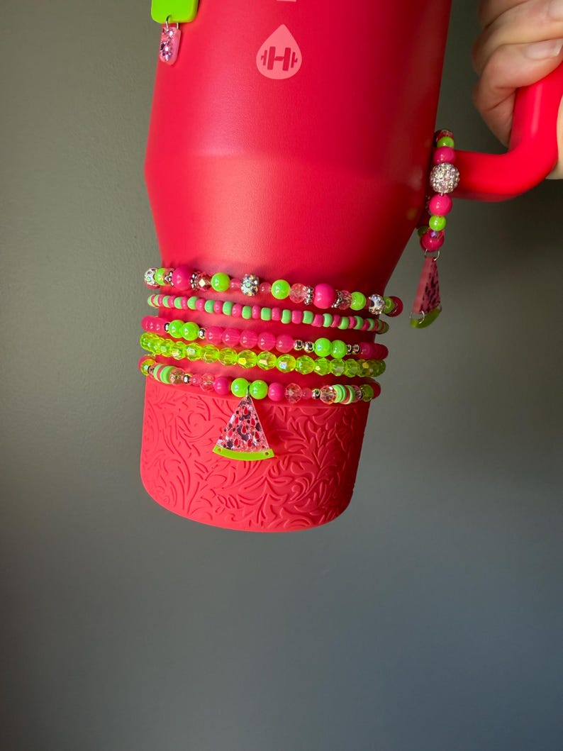 May include: A bright pink tumbler with a textured base and a handle, adorned with multiple stacked beaded bracelets in neon green and pink. The bracelets feature various round beads, faceted crystals, and a dangling watermelon slice charm.