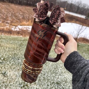 May include: A brown and gold stainless steel tumbler with a zebra print design. The tumbler has a black handle and a brown glitter bow on top. The tumbler is decorated with gold and brown beads around the bottom.