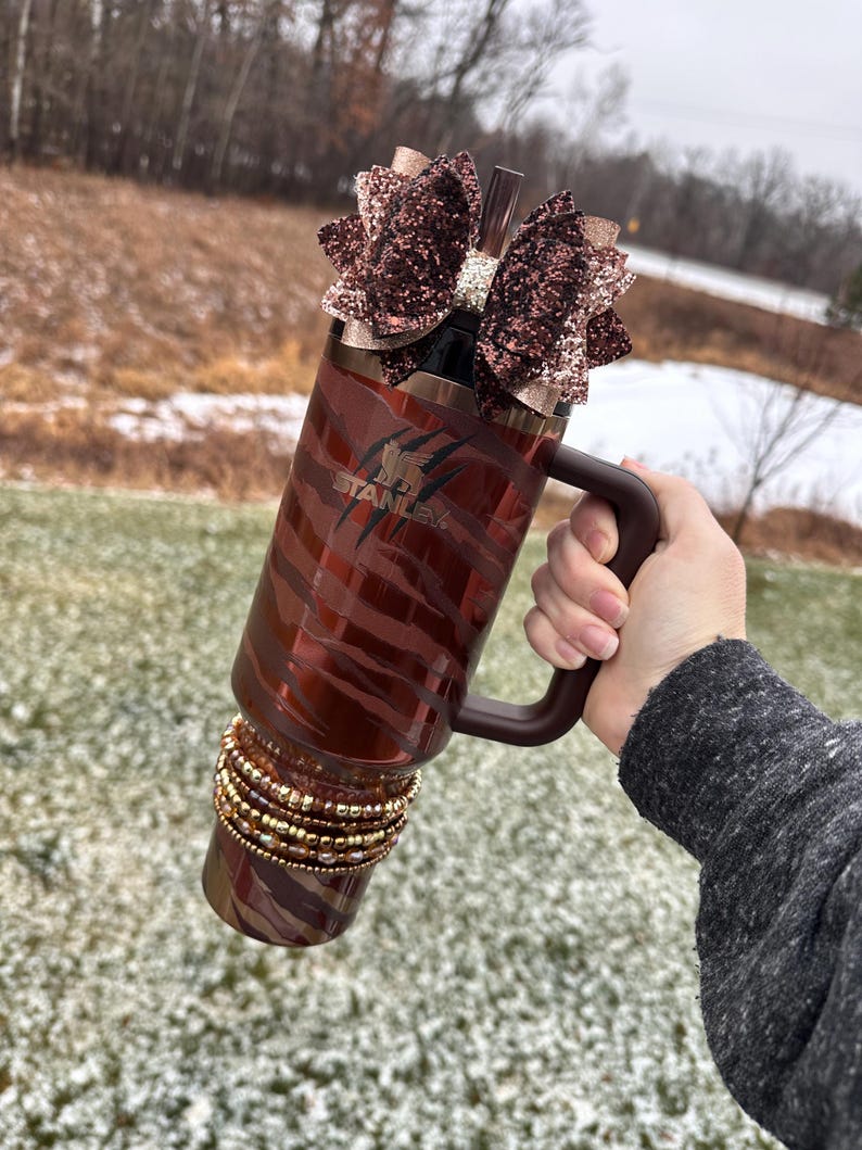 May include: A brown and gold stainless steel tumbler with a zebra print design. The tumbler has a black handle and a brown glitter bow on top. The tumbler is decorated with gold and brown beads around the bottom.