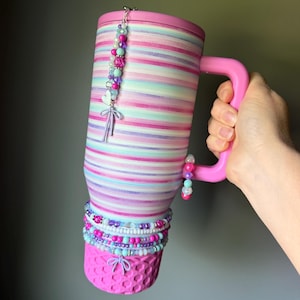 Op de afbeelding: Een hoge ge&iuml;soleerde beker met een roze deksel en handvat, voorzien van horizontale strepen in tinten roze, paars en lichtblauw. De beker is versierd met meerdere kralenbedels, waaronder een hartje en een strik, en een roze getextureerde basis.