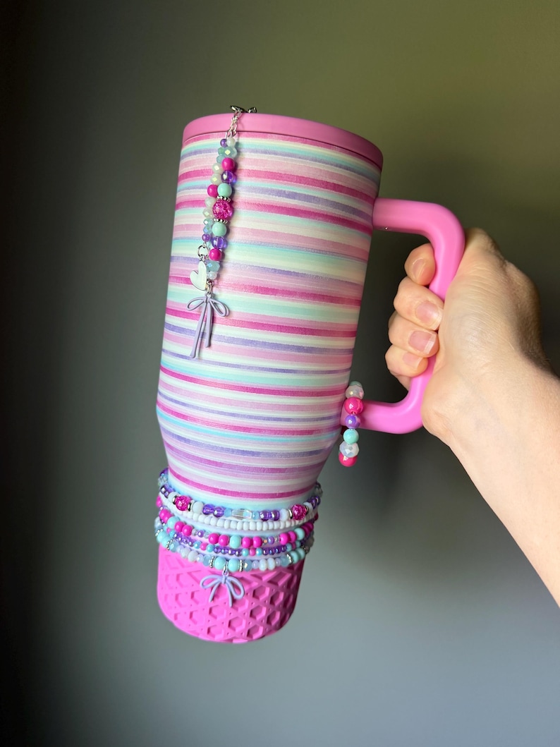 Op de afbeelding: Een hoge ge&iuml;soleerde beker met een roze deksel en handvat, voorzien van horizontale strepen in tinten roze, paars en lichtblauw. De beker is versierd met meerdere kralenbedels, waaronder een hartje en een strik, en een roze getextureerde basis.