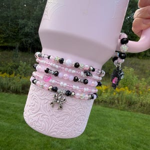 May include: A light pink tumbler with a textured spiderweb pattern, adorned with a stack of beaded bracelets featuring black, white, and pink beads, a silver spider charm, a pink heart bead, and a dangling black spider charm on the handle.