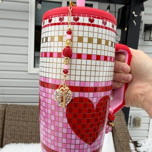 May include: A large, mosaic-style tumbler with a red lid and handle. The tumbler features a pattern of pink, white, and gold squares, with a large red heart design. A charm with a heart and key hangs from the lid. The tumbler is adorned with beaded accents.