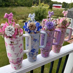 K&ouml;nnte beinhalten: Vier Edelstahlbecher mit Blumenmustern und verschiedenen Glitzerakzenten. Jeder Becher hat einen wei&szlig;en Griff und einen Strohhalm mit einer dekorativen Schleife oben. Die Becher sind in einer Reihe auf einem wei&szlig;en Gel&auml;nder angeordnet.