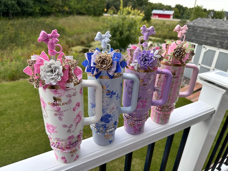 K&ouml;nnte beinhalten: Vier Edelstahlbecher mit Blumenmustern und verschiedenen Glitzerakzenten. Jeder Becher hat einen wei&szlig;en Griff und einen Strohhalm mit einer dekorativen Schleife oben. Die Becher sind in einer Reihe auf einem wei&szlig;en Gel&auml;nder angeordnet.