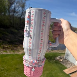 May include: A white tumbler with a floral and striped pattern, adorned with pink and blue beads and charms. The bottom has a pink knitted sleeve. A beaded charm hangs from the lid. The tumbler is held by a person.