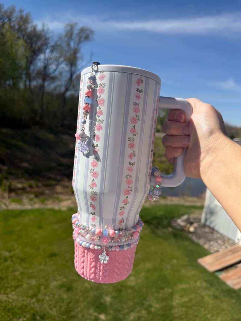 May include: A white tumbler with a floral and striped pattern, adorned with pink and blue beads and charms. The bottom has a pink knitted sleeve. A beaded charm hangs from the lid. The tumbler is held by a person.