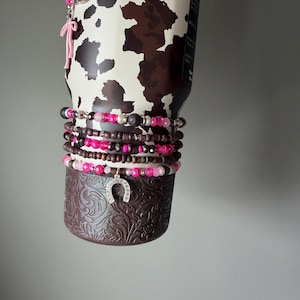 May include: A stainless steel tumbler with a brown and white cow print pattern, adorned with a stack of beaded bracelets featuring pink, brown, and white beads, a silver horseshoe charm, and a dangling pink bow and boot charm accessory.