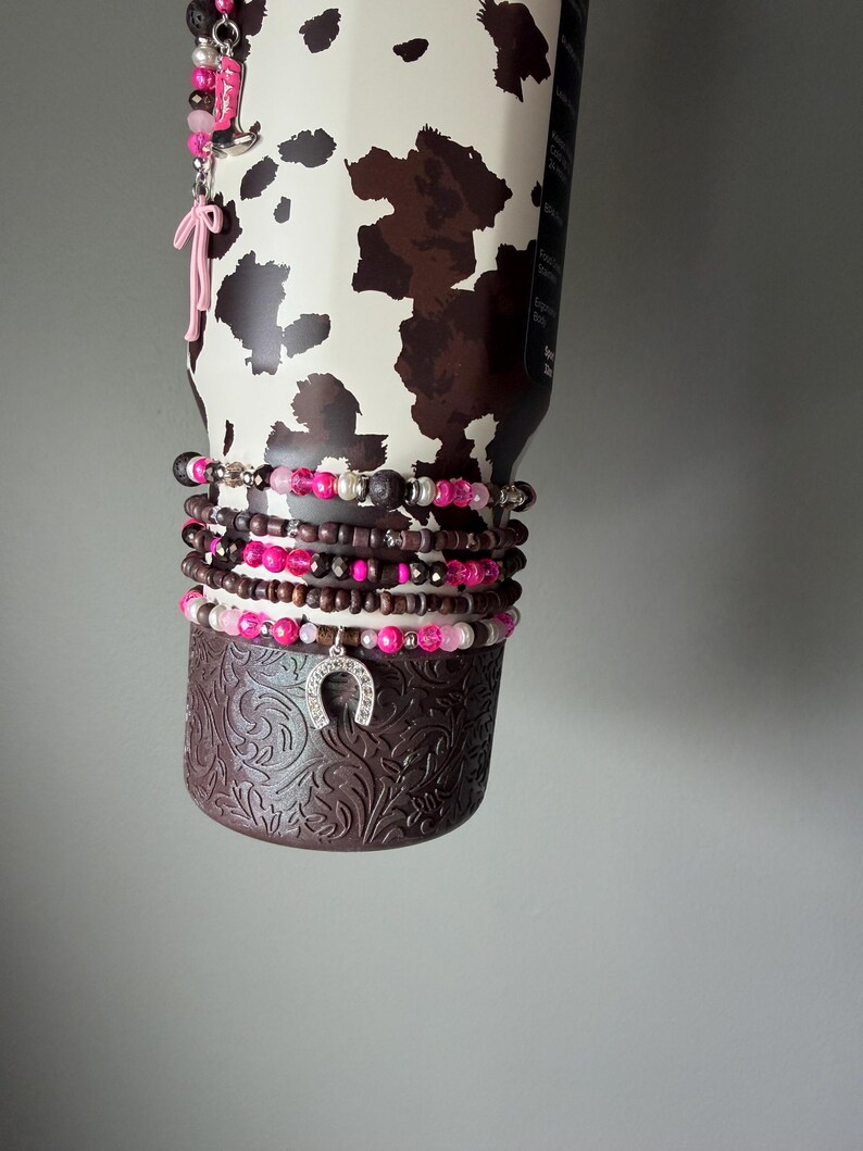 May include: A stainless steel tumbler with a brown and white cow print pattern, adorned with a stack of beaded bracelets featuring pink, brown, and white beads, a silver horseshoe charm, and a dangling pink bow and boot charm accessory.