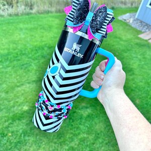 May include: A black and white striped stainless steel water bottle with a blue handle. The bottle has a black and white bow with pink and blue glitter on top. The bottle is decorated with black and white beaded bracelets.