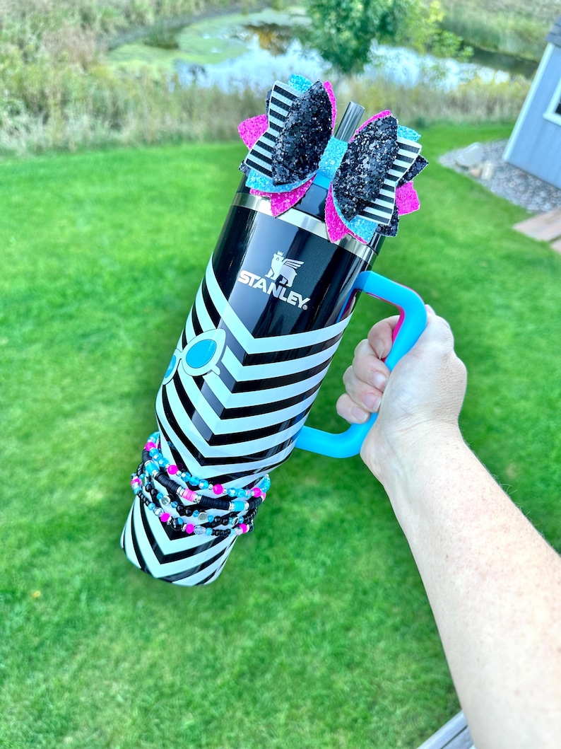 May include: A black and white striped stainless steel water bottle with a blue handle. The bottle has a black and white bow with pink and blue glitter on top. The bottle is decorated with black and white beaded bracelets.